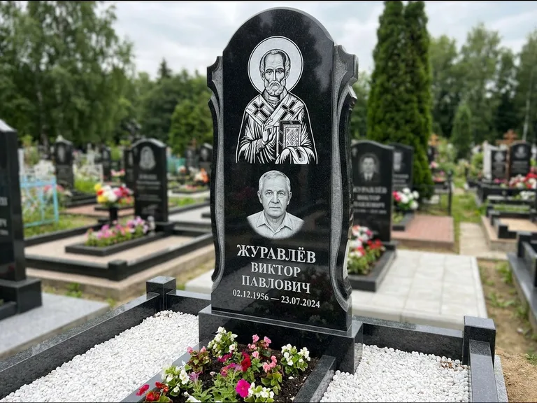 Памятник с гравировкой иконы: стела с образом в полный рост на обороте — гравировка образа Матроны Московской, низкие кусты спиреи в цвету, работа «Лебедева» в Кемерово